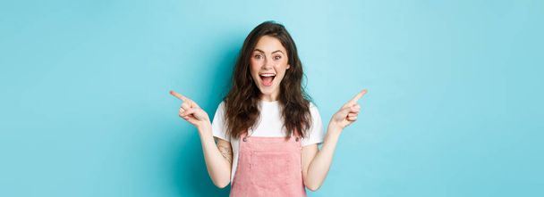 Pick your side. Smiling excited woman screaming of joy, pointing fingers sideways at left and right copy space, showing advertisement good deal, standing against blue background. - Foto, Bild