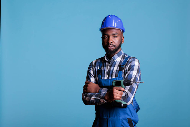 African american builder in coverall protective helmet hold electric drill isolated on blue background studio. Serious builder looks confident holding work tools while looking at the camera. - 写真・画像