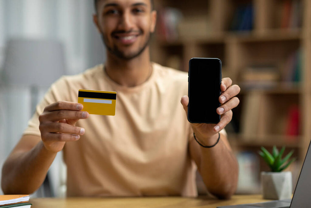 Happy young arab man showing smartphone with blank screen and credit card, selective focus, cropped, mockup. Sale, online shopping, application for purchases, finance and banking with new device - Foto, Imagen