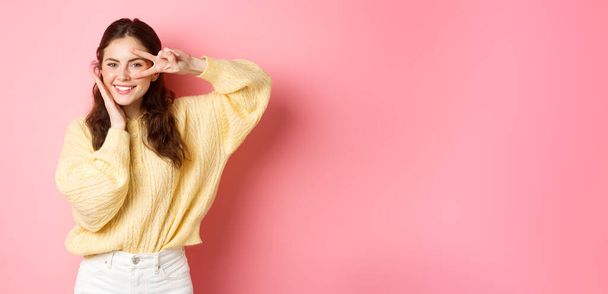 Aantrekkelijk brunette meisje in casual kleding, aanraken van haar perfecte schone gezicht, tonen vrede v-teken op oog, staan tegen roze achtergrond. - Foto, afbeelding