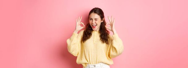 Portrait of cheerful pretty girl winking and saying yes, showing okay sign, alright gesture, praise good job, well done, standing pleased against pink background. - Foto, immagini