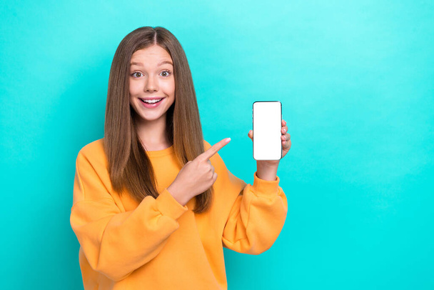 Portrait photo of excited cute girl wear orange sweater hold phone finger directing mockup app for ebooks education isolated on cyan color background. - Фото, изображение