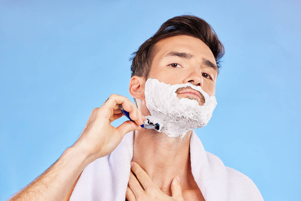Portrait, shave and bathroom with a man model shaving his face in studio on a blue background for grooming. Beauty, skincare and shaving foam with a handsome young male using a razor in the morning. - Foto, Imagen