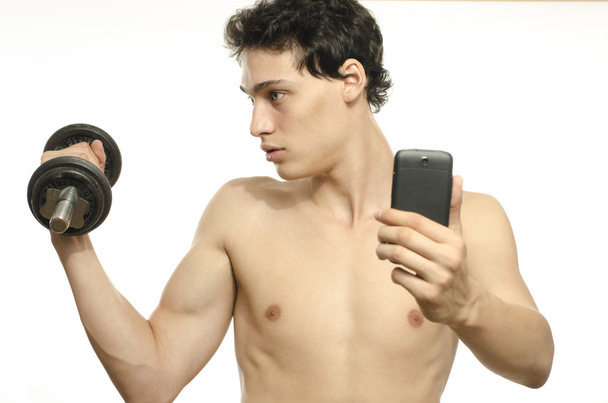 Skinny man taking a selfie with his phone while training his bicep muscle. Beautiful teenager lifting a dumbbell and taking a photo for facebook.Anorexic young man training to become stronger - Foto, Imagem