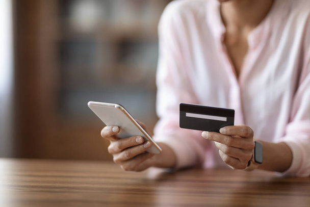 Unerkennbare Frau in pinkfarbener Bluse sitzt am Schreibtisch, hält moderne Handy- und Plastikkreditkarten in der Hand, kauft online ein oder bezahlt Rechnungen im Internet, beschnitten, kopiert. Online-Banking-Konzept - Foto, Bild