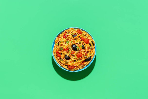 Above view with a plate of pasta puttanesca, minimalist on a green background. Traditional italian pasta dish, with tomato sauce, black olives and capers. - Photo, Image