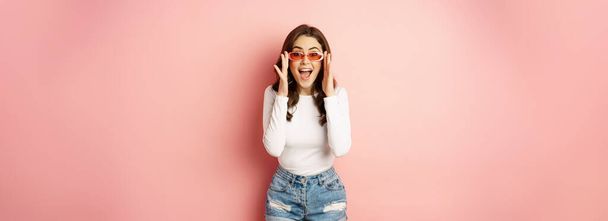 Enthusiastic brunette girl in stylish sunglasses, reacts surprised and amazed, looking with disbelief at camera, standing over pink background. - 写真・画像