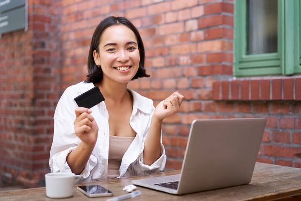 Hermosa mujer asiática joven con tarjeta de crédito, sentado cerca de la computadora portátil y sonriendo, el pago de facturas, tiendas en línea, orden smth en el ordenador. - Foto, imagen