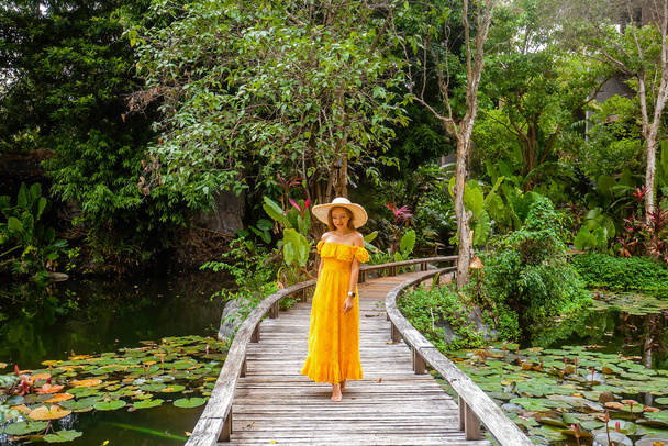 Happy woman in yellow dress enjoy her summer vacation in resort. Female traveller walking on wooden bridge among tropical greenery and green pond with water lilies in hot sunny day. Holiday and travel - Fotografie, Obrázek