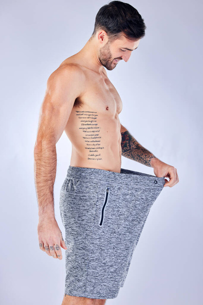 Motivation, diet and weight loss, man checking pants size change from workout and healthy lifestyle on studio background. Fitness, nutrition and wellness, balance in body care, male model with tattoo. - Foto, Bild