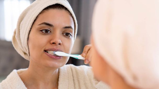 Гігієна ротової порожнини, здорові зуби та догляд. Молода жінка чистить зуби зубною щіткою і дивиться у дзеркало в інтер'єрі ванної
 - Фото, зображення
