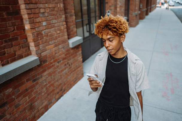Joven hombre multirracial positivo en ropa elegante sosteniendo el teléfono móvil inteligente mientras camina al aire libre y en busca de dirección con aplicación para teléfonos inteligentes en la calle de la ciudad - Foto, imagen