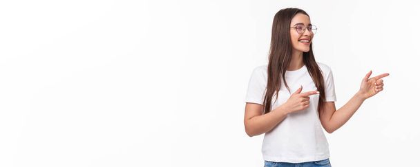 Waist-up portrait of cheerful young female student, programmer, pointing fingers and looking at right copy space, smiling and laughing as seeing something entertaining and funny, white background. - Foto, imagen