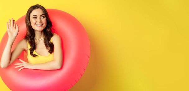 Ευτυχισμένη νεαρή γυναίκα φορώντας ροζ δαχτυλίδι κολύμβησης, κουνώντας το χέρι και λέγοντας γεια, απολαμβάνοντας τις διακοπές, καλοκαιρινές διακοπές στην παραλία, στέκεται πάνω από κίτρινο φόντο. - Φωτογραφία, εικόνα
