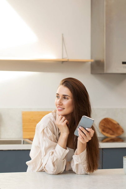 Μια ελκυστική μελαχρινή κρατά ένα τηλέφωνο στο χέρι της και χαμογελώντας κοιτάζει από το πλάι, ακουμπώντας στον πάγκο στην κουζίνα. Η έννοια των online αγορών, παραγγελία τροφίμων στο σπίτι. - Φωτογραφία, εικόνα