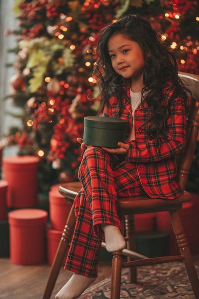 Happy japan little girl dreamer waiting miracle from Santa posing on floor near noel tree. Asian kid in red plaid outfit celebrating new year with gifts boxes at bokeh xmas lights eve 25 december - Photo, Image