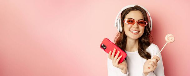 Stylová mladá žena s lízátkem a mobilem, nosí sluneční brýle a sluchátka, poslouchá hudbu, stojí nad růžovým pozadím. - Fotografie, Obrázek