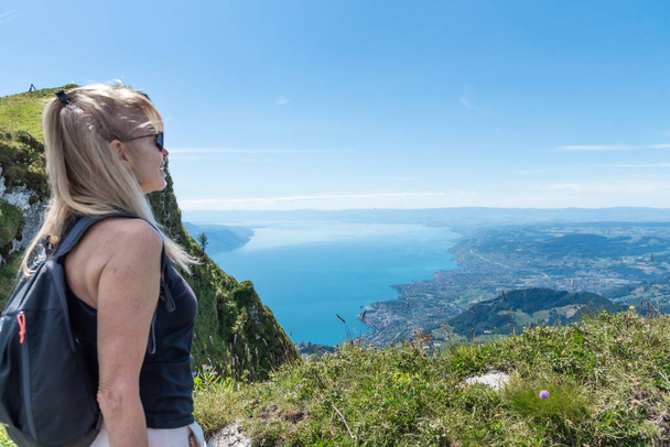 Ξανθιά ενήλικη γυναίκα με γυαλιά ηλίου σακίδιο κοιτάζοντας από την άποψη του βουνού στη λίμνη της Γενεύης με τις πόλεις Montreux και Vevey - Φωτογραφία, εικόνα
