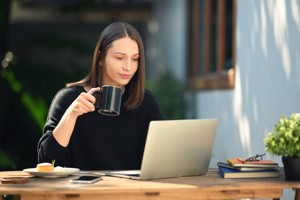 Beautiful young woman drinking hot coffee and working with laptop while sitting at the table in outdoor coffee shop. - Foto, imagen