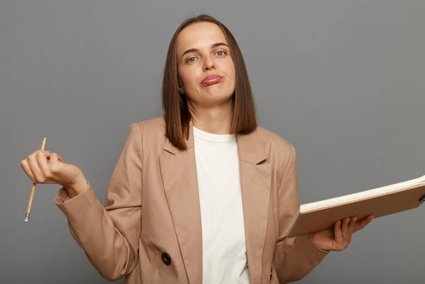 Horizontal shot of woman wearing beige jacket standing with pencil and clipboard posing isolated on gray background, being sad and upset, looking at camera with pout lips and shrugging shoulders. - Photo, Image
