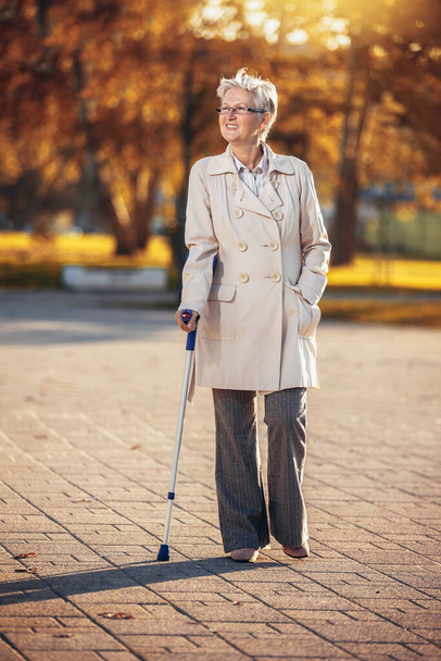Oudere vrouw met kruk op een wandeling in het najaarspark. - Foto, afbeelding
