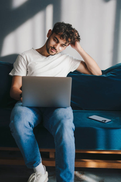 Nyugodt fiatalember, aki a laptop számítógépén dolgozik, kék kanapén ül, és otthon tölti az idejét a nappaliban. nternet kommunikáció. Modern kommunikáció - Fotó, kép