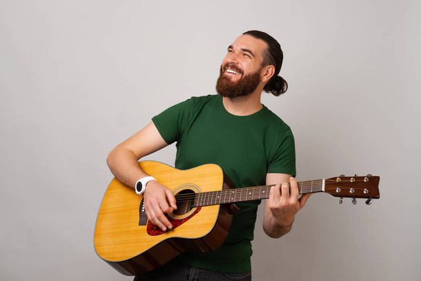 Hombre fuerte guapo está tocando una guitarra acústica mientras mira hacia arriba y sonríe. Estudio sobre fondo gris. - Foto, imagen
