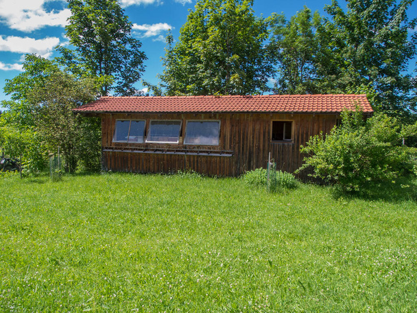 Frontal view of the long side with three glass windows of a beekeeper's house in Bavaria on a green meadow with group of trees in the background under a blue sky with a few white clouds. - Fotó, kép