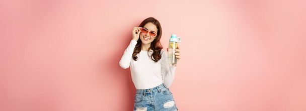 Stylish girl in spring outfit, wearing sunglasses, holding water bottle with lemon, healthy drink, laughing and smiling, standing over pink background. - Фото, изображение