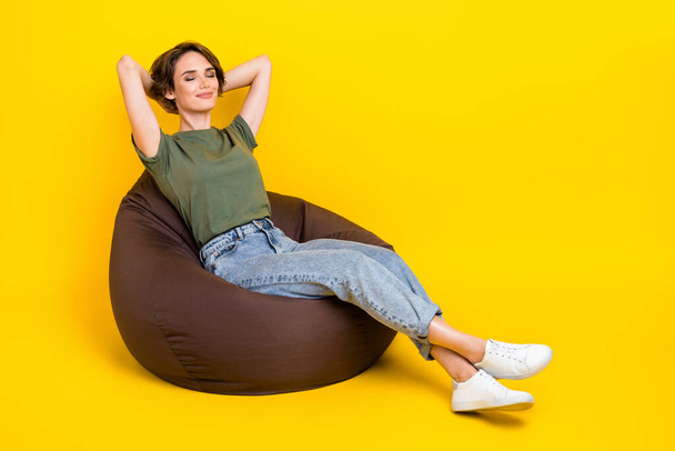Full size photo of cheerful calm girl bob hairstyle wear khaki t-shirt sit on pouf hands behind head isolated on yellow color background. - 写真・画像