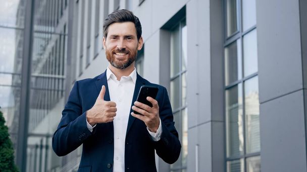 Dışarıda duran mutlu bir adam, elinde telefonla gülümseyen bir işadamı iş için yeni mobil uygulamalar kullanıyor. Onaylama jesti gösteriyor. Mükemmel bir sonuç için en iyi kalite öneriliyor. - Fotoğraf, Görsel