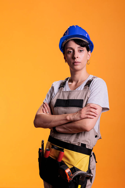 Portrait of female construction worker posing with arms crossed in studio. Professional woman in construction industry looking confident, standing in renovation overalls over yellow background. - Photo, Image