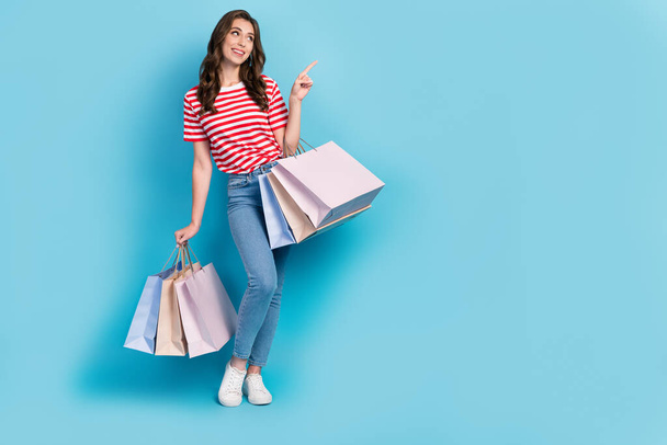 Full length photo of pretty dreamy girl dressed red t-shirt pointing empty space holding shoppers isolated blue color background. - 写真・画像