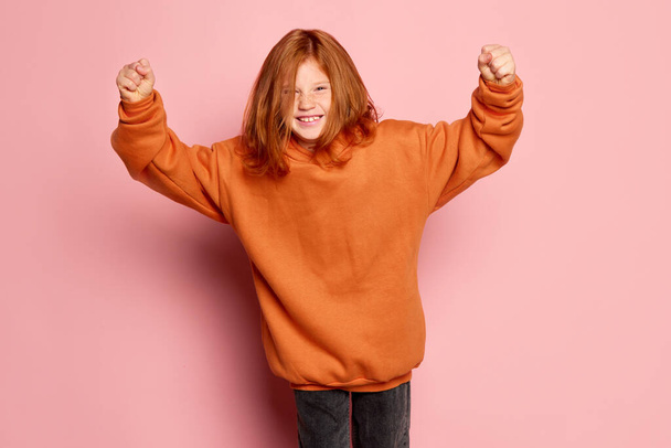 Expressive teen girl, modern kid with long red hair and freckles looking at camera isolated over pink background. Concept of children emotions, casual sports fashion style, facial expressions. - 写真・画像