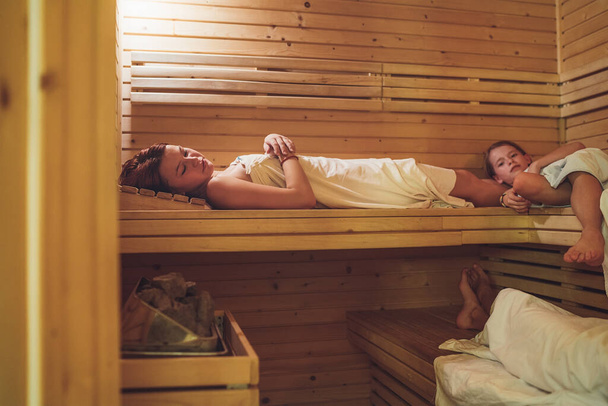 Relaxing kids sisters girls wrapped in white sheets lying and sweating on wooden benches in hot Finnish sauna enjoying pleasant body care temperature treatment. Healthy people lifestyle concept. - Fotografie, Obrázek