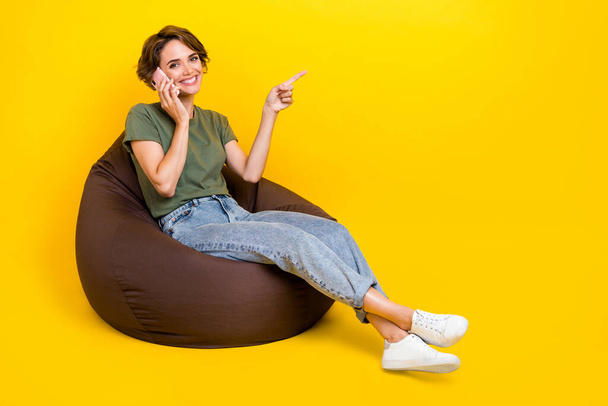 Full length photo of nice positive woman brown hair talking phone sit on pouf directing empty space isolated on yellow color background. - Valokuva, kuva