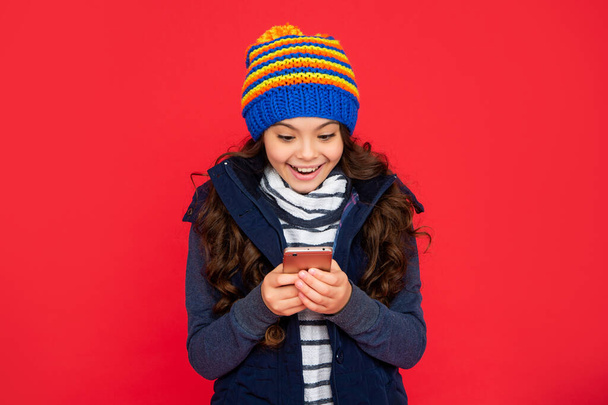 portrait of child blogging with phone. express positive emotion. greeting with winter holidays. smiling amazed kid chatting online in hat on red background. teen girl texting on smartphone. - Photo, image