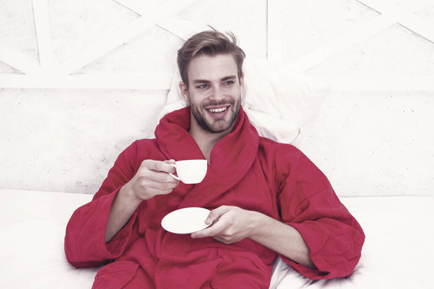 Drinking coffee for breakfast. Happy guy drinking coffee in bed. Bearded man in bathrobe holding drinking cup and smiling. Enjoying drinking tea or other hot beverage after bath spa. - Фото, зображення