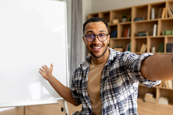Vrolijke Arabische mentor man glimlachend naar de camera en wijzend naar whiteboard, met online les in de klas. Onderwijsconcept op afstand - Foto, afbeelding