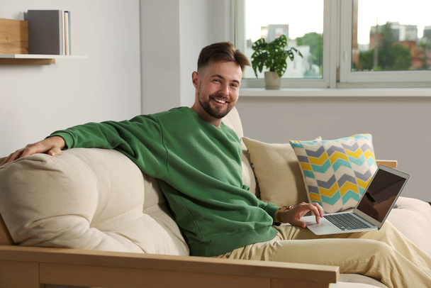 Handsome man with laptop sitting on sofa at home - Fotoğraf, Görsel