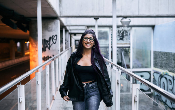 young latina woman glasses brunette fur jacket big black boots with a big smile happy and content walking down the glass walkway - Urban concept - Photo, image