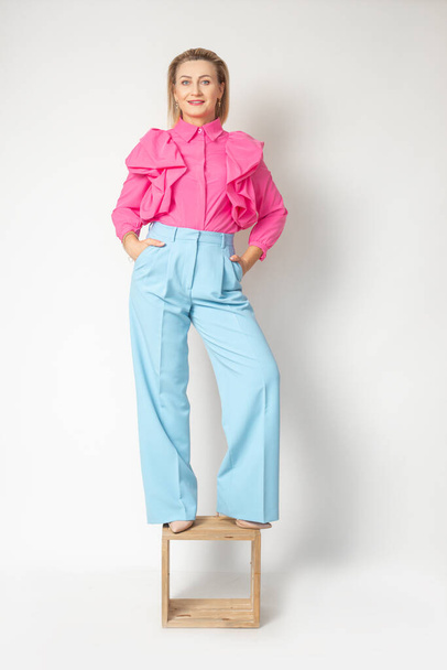 Χαριτωμένη ενήλικη γυναίκα με ροζ μπλούζα και μπλε παντελόνι που στέκεται στην καρέκλα, απομονωμένη σε λευκό. Full length φωτογραφία της αυτοπεποίθηση επιτυχημένη επιχειρηματίας. Όμορφοι άνθρωποι, ρούχα μόδας. - Φωτογραφία, εικόνα