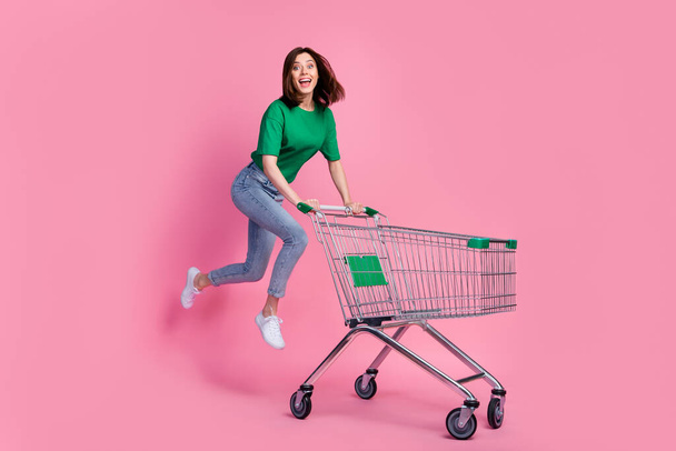 Full body photo of carefree overjoyed lady jumping push supermarket trolley isolated on pink color background. - 写真・画像