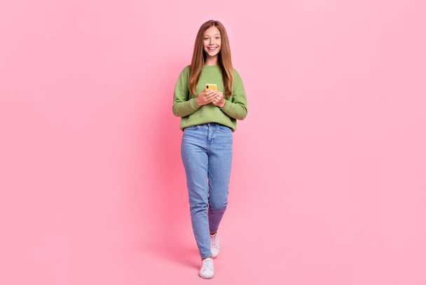 Full length photo of cute cheerful school girl dressed green sweatshirt communicating modern device isolated pink color background. - Foto, Imagen