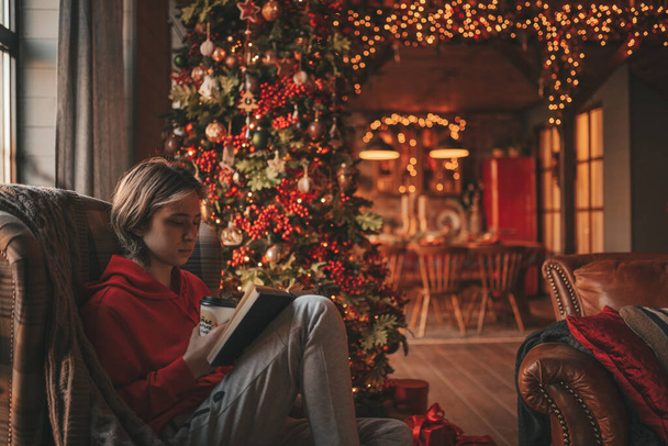 Young teen boy with long hair holding and reading study book for distance learning at home in holidays. Stylish zoomer gen Z celebrates new year with xmas tree bokeh lights garlands eve 25 december - Foto, Imagen