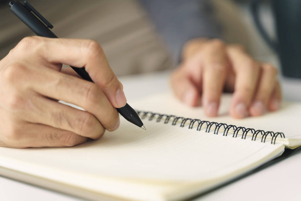 Крупный план молодого человека в повседневной ткани руки, записанные на блокноте, ноутбук с шариковой ручкой на столе. - Фото, изображение
