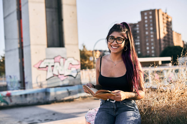 junge lateinische brünette Frau mit Brille, Lippenpiercing, Jeans und langen Haaren, die in einem Stadtpark sitzt und mit einem Buch in der Hand in die Kamera lächelt - Urban concept - Foto, Bild