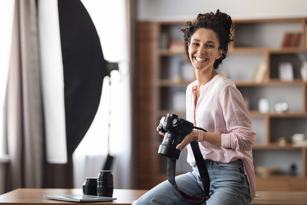 Stylová kreativní brunetka atraktivní mladá žena v ležérní profesionální fotograf nebo blogger sedí u stolu doma studio nebo domácí kancelář drží fotoaparát a usmívá se uvnitř, kopírovat prostor - Fotografie, Obrázek