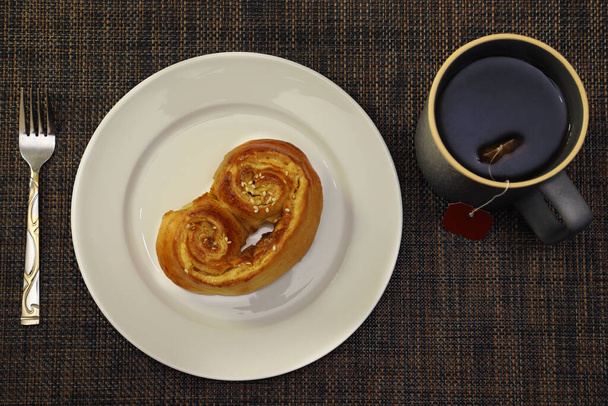 Rollo con canela y semillas de sésamo en un plato blanco con una taza de té. Vista desde arriba - Foto, Imagen