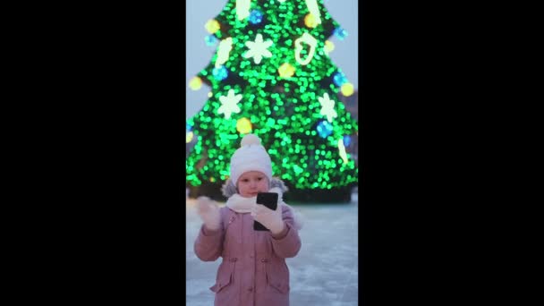 Ein kleines Mädchen steht auf der Straße in der Nähe des Weihnachtsbaums, telefoniert mit dem Smartphone und schickt Glückwünsche. Online-Glückwünsche per Telefon - Filmmaterial, Video